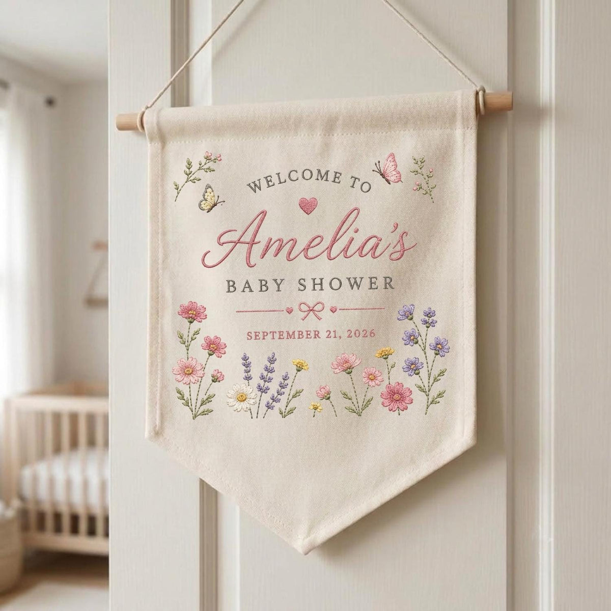 May include: A cream-colored fabric wall banner hanging from a wooden dowel with string. The banner features the text "WELCOME TO Amelia's BABY SHOWER SEPTEMBER 21, 2026" in pink and gray, decorated with colorful floral illustrations and butterflies.