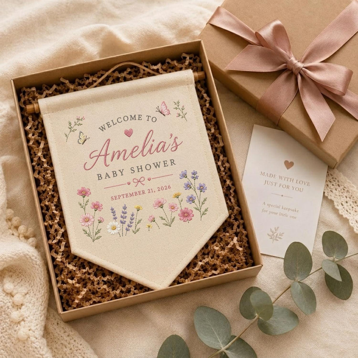 May include: A cream-colored fabric banner with wooden dowels, featuring the text 'WELCOME TO Amelia's BABY SHOWER SEPTEMBER 21, 2026' and floral illustrations, presented in a gift box with a matching card and a satin ribbon bow.