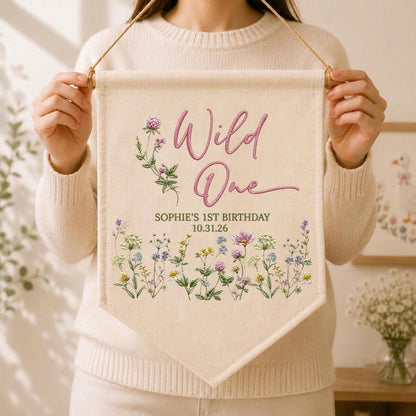 May include: A cream-colored fabric wall banner featuring the pink text Wild One, the name Sophie's 1st Birthday, and the date 10.31.26. The design includes printed illustrations of various colorful wildflowers and green stems.