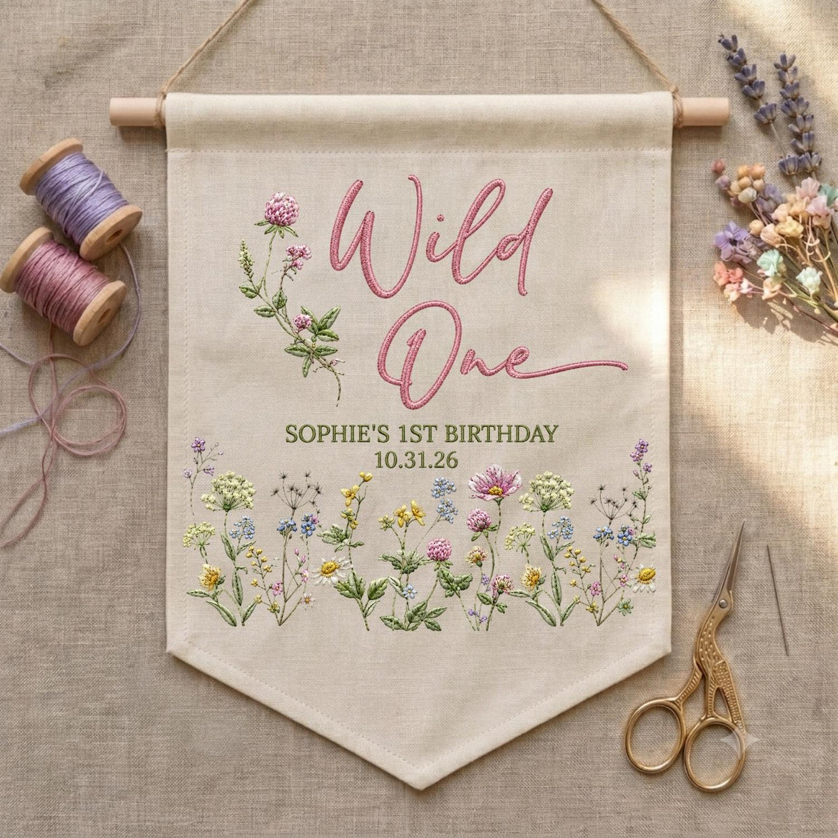 May include: A beige fabric wall banner featuring embroidered pink text that reads Wild One, with the text Sophie's 1st Birthday 10.31.26 below. The banner is decorated with embroidered wildflowers in pink, yellow, blue, and green hues.
