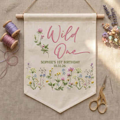 May include: A beige fabric wall banner featuring embroidered pink text that reads Wild One, with the text Sophie's 1st Birthday 10.31.26 below. The banner is decorated with embroidered wildflowers in pink, yellow, blue, and green hues.