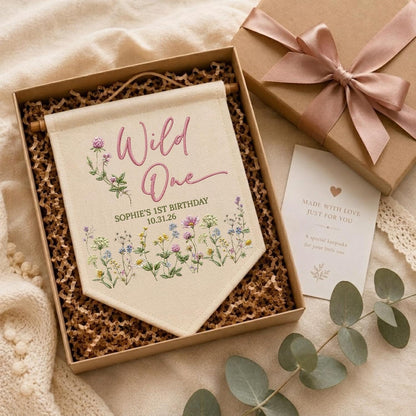 May include: A cream-colored fabric wall banner featuring the pink text Wild One, the name Sophie's 1st Birthday, and the date 10.31.26, decorated with colorful wildflower illustrations, presented in a gift box with a matching card and ribbon.