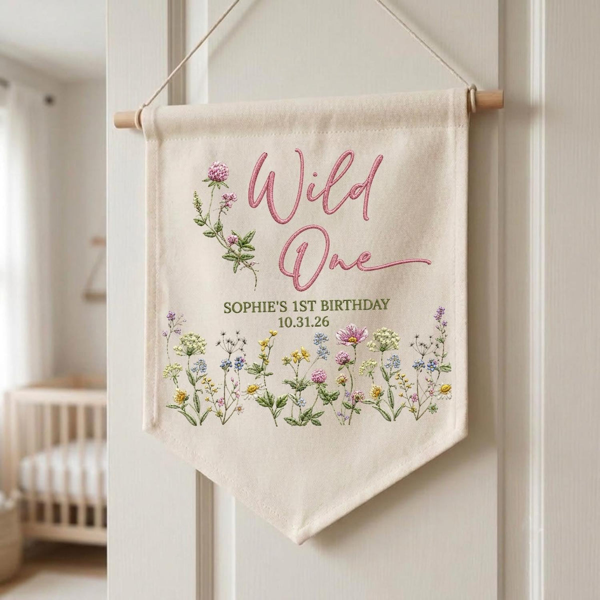 May include: A cream-colored fabric wall banner featuring the pink embroidered text Wild One, followed by SOPHIE'S 1ST BIRTHDAY and 10.31.26. The banner is decorated with a colorful botanical illustration of various wildflowers and clover.