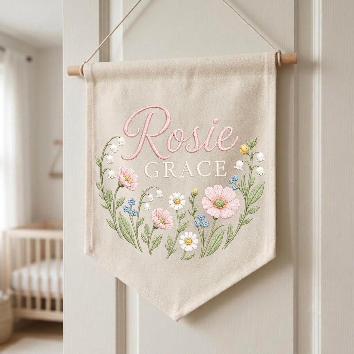 May include: A beige fabric wall banner hanging from a wooden dowel with a string. The banner features the name Rosie Grace in pink and white script and block lettering, surrounded by a colorful illustration of various wildflowers and greenery.