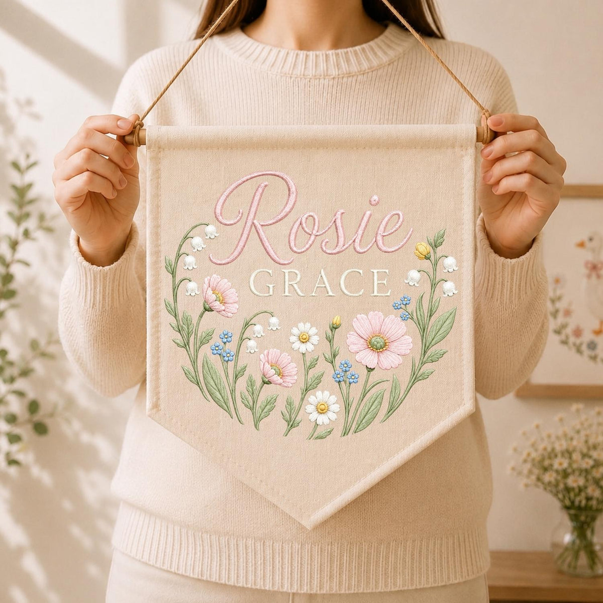 May include: A beige fabric wall banner featuring the name Rosie Grace in pink and white script and block lettering. The design includes a floral wreath with pink cosmos, white daisies, blue forget-me-nots, and lily of the valley stems.