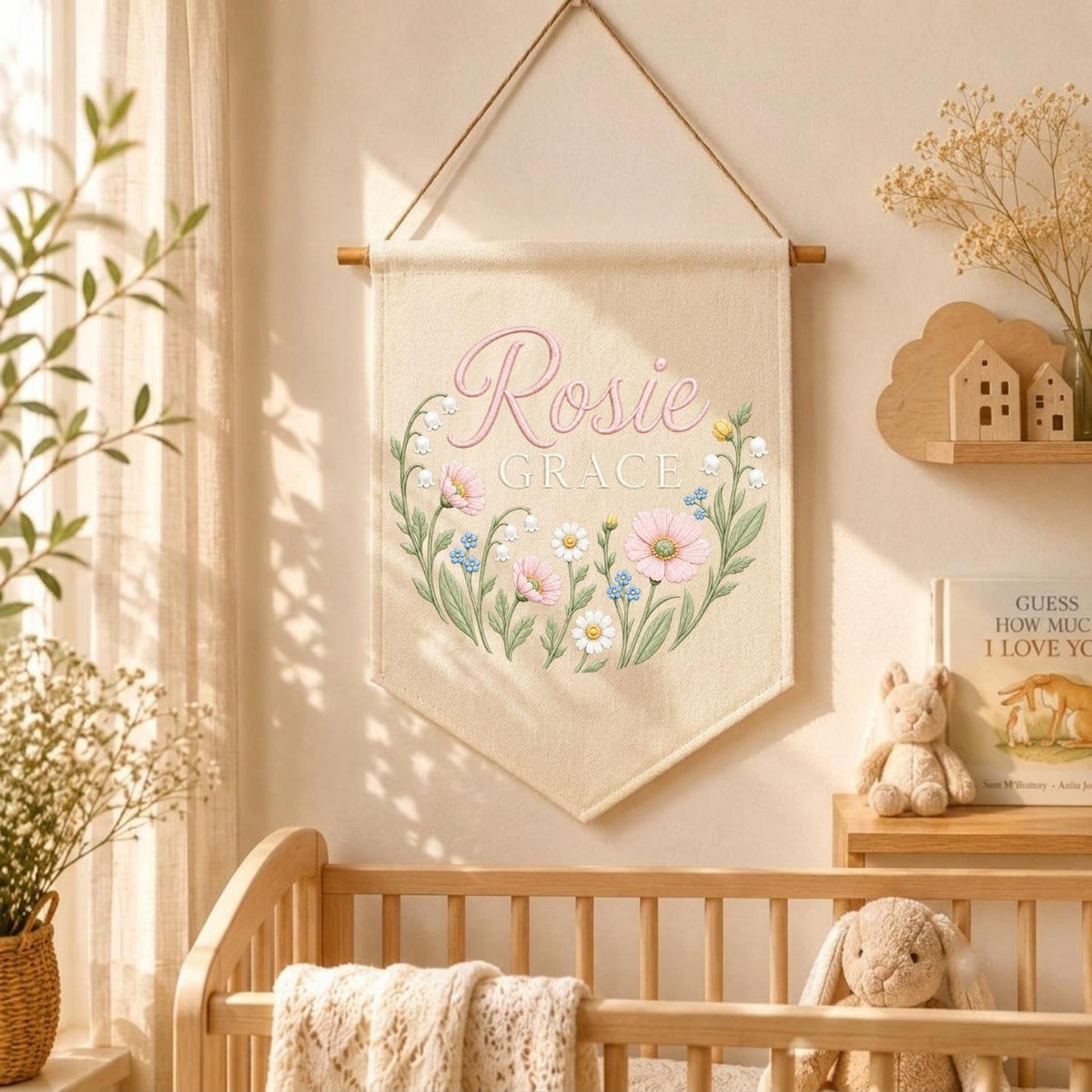 May include: A cream-colored fabric wall banner hanging from a wooden rod with a string. The banner features the name Rosie Grace in pink script and block lettering, surrounded by a circular illustration of pink and white wildflowers.