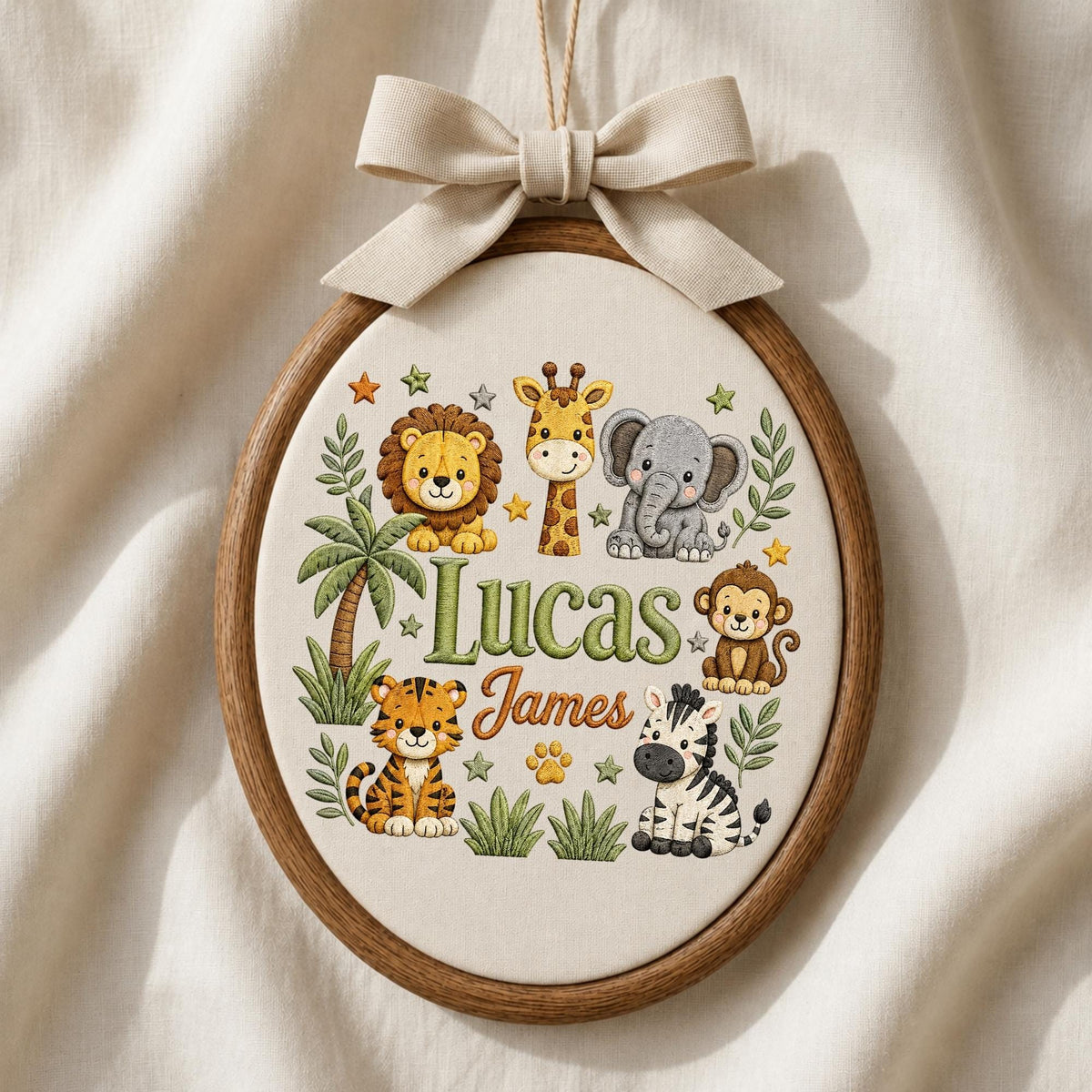 May include: An oval wooden embroidery hoop containing fabric with a stitched jungle theme. The design features a lion, giraffe, elephant, monkey, tiger, and zebra, with the names Lucas and James in the center, topped with a beige fabric bow.