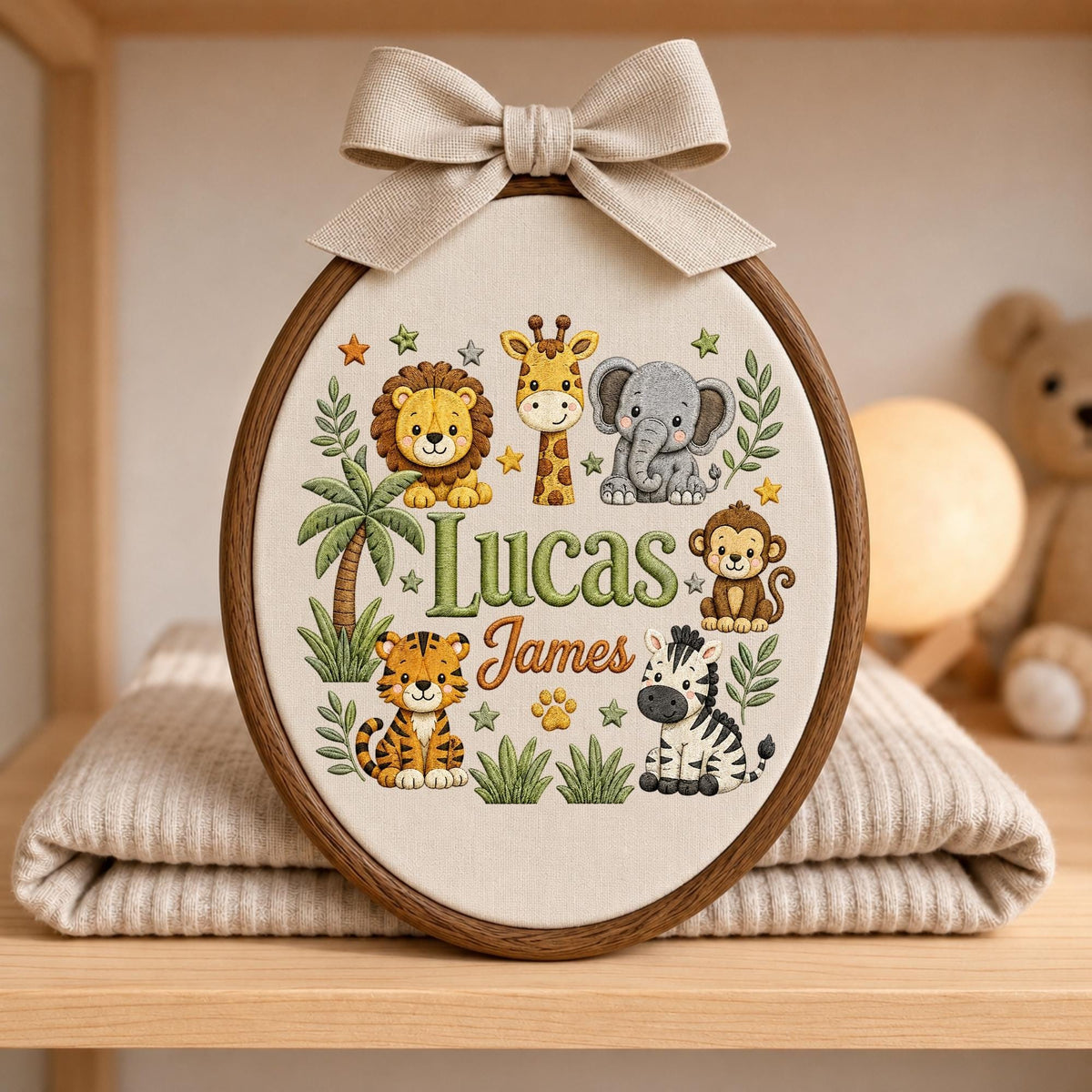 May include: An oval wooden embroidery hoop containing fabric with a stitched jungle animal design featuring a lion, giraffe, elephant, monkey, tiger, and zebra. The names Lucas and James are embroidered in the center, topped with a beige fabric bow.