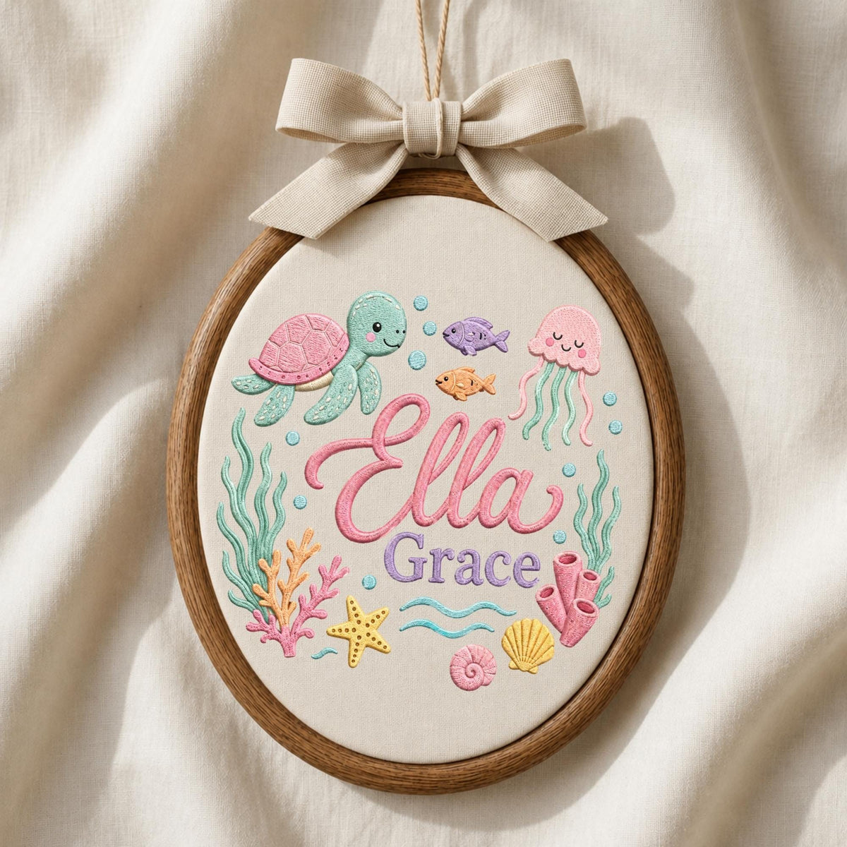 May include: An oval embroidery hoop featuring a cream fabric background with colorful stitched sea life including a turtle, jellyfish, fish, starfish, and coral. The name Ella Grace is embroidered in pink and purple script in the center.