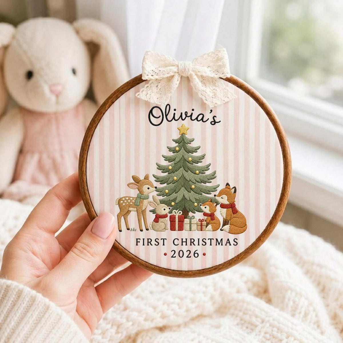May include: A circular wooden embroidery hoop frame containing fabric with a pink and white striped pattern. The design features a Christmas tree, a deer, a rabbit, and two foxes with gifts. Text reads Olivia's FIRST CHRISTMAS 2026.