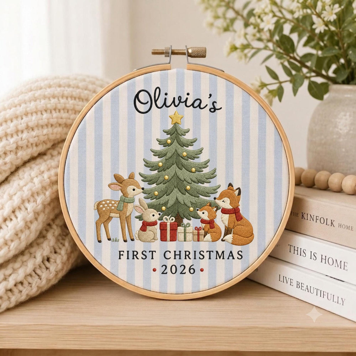 May include: A round wooden embroidery hoop containing fabric with light blue vertical stripes. The design features a green Christmas tree, a deer, a rabbit, and a fox with gifts. Text reads: Olivia's FIRST CHRISTMAS 2026.