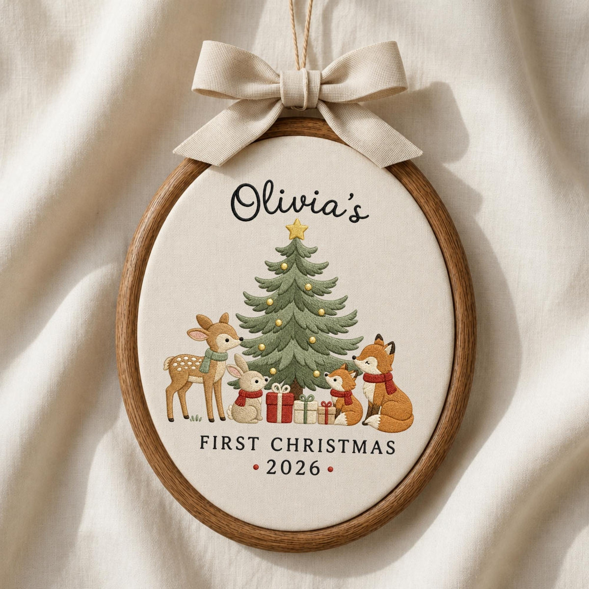 May include: An oval wooden embroidery hoop ornament featuring a cream fabric center with a printed Christmas tree, a deer, a rabbit, and two foxes. The text reads Olivia's FIRST CHRISTMAS 2026. A beige fabric bow is attached to the top of the hoop.