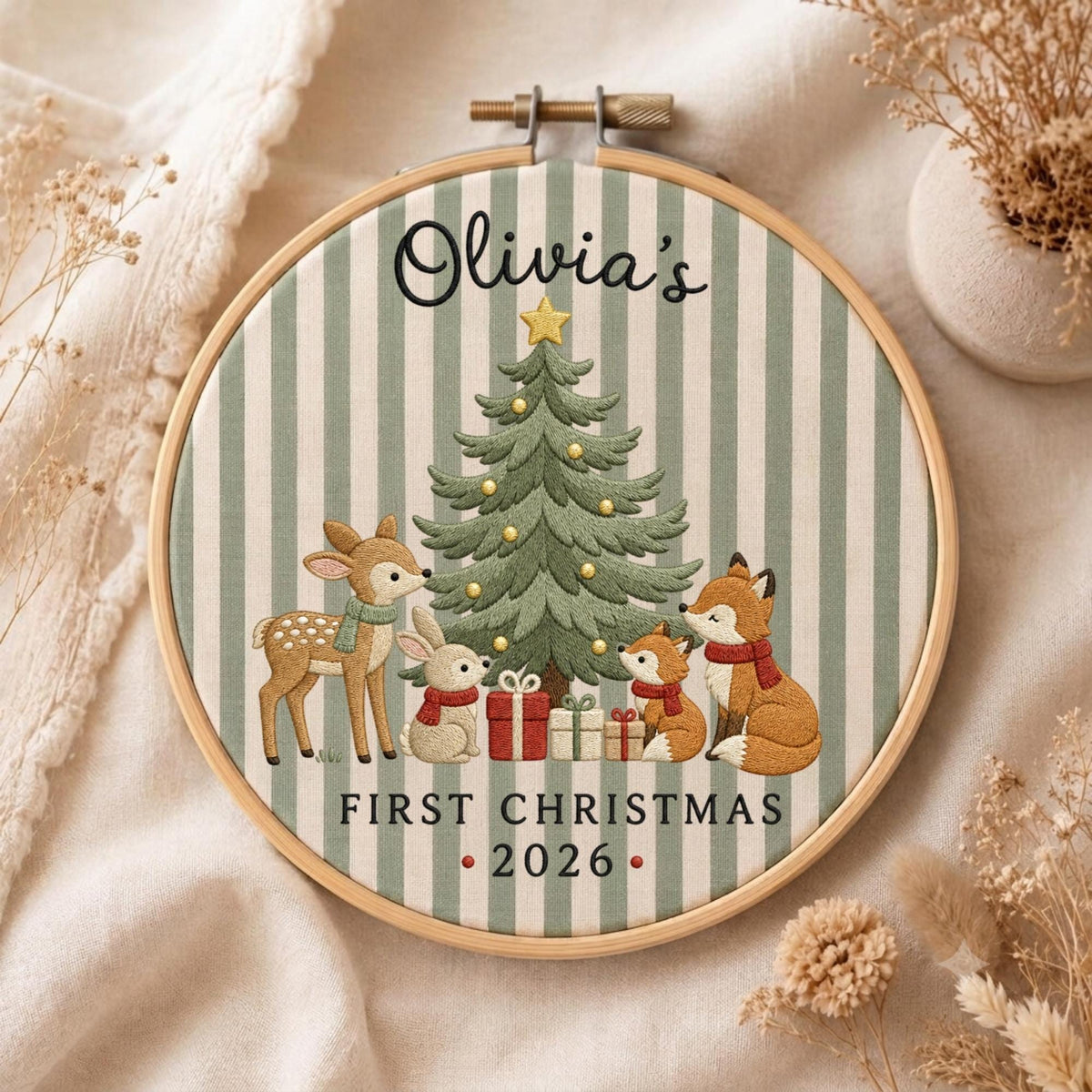 May include: A round wooden embroidery hoop containing fabric with green and white vertical stripes. The embroidery features a Christmas tree, a deer, a rabbit, and a fox with gifts. Text reads: Olivia's FIRST CHRISTMAS 2026.