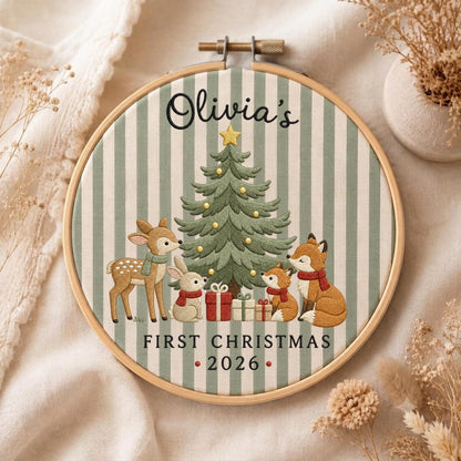 May include: A round wooden embroidery hoop containing fabric with green and white vertical stripes. The embroidery features a Christmas tree, a deer, a rabbit, and a fox with gifts. Text reads: Olivia's FIRST CHRISTMAS 2026.