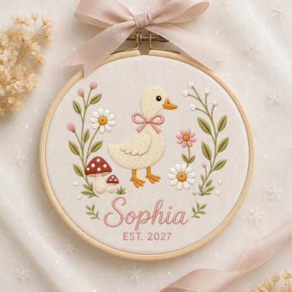 May include: A circular wooden embroidery hoop containing cream fabric with a stitched design of a light yellow duckling, pink flowers, white daisies, and red mushrooms. The name Sophia and the text EST. 2027 are embroidered in pink below the duck.