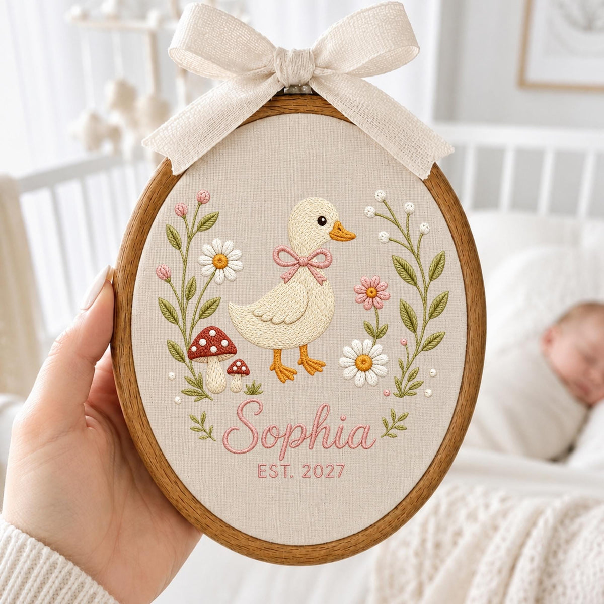 May include: An oval wooden embroidery hoop containing a cream-colored fabric with a stitched design of a duckling, mushrooms, and flowers. The name Sophia and the text EST. 2027 are embroidered in pink. A beige ribbon bow is tied at the top.