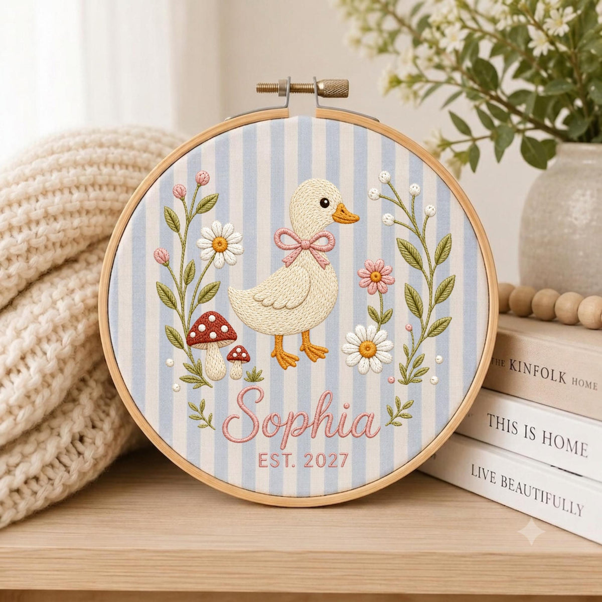 May include: A circular wooden embroidery hoop containing fabric with light blue and white vertical stripes. The embroidery features a central cream-colored duckling with a pink bow, surrounded by daisies, mushrooms, and the text Sophia EST. 2027.