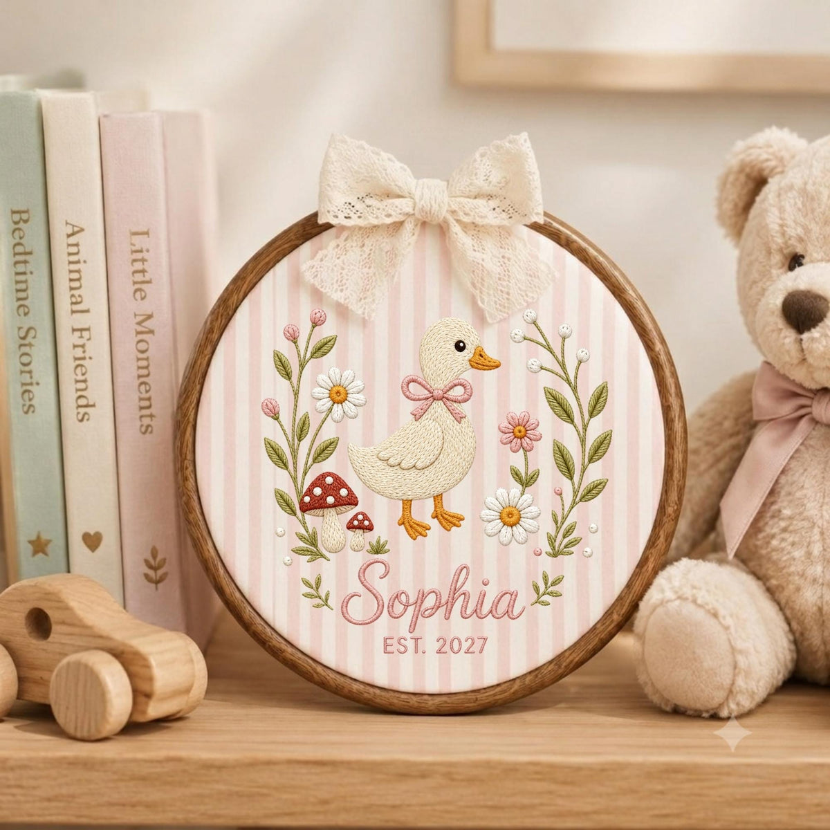 May include: A circular wooden embroidery hoop featuring a light pink and white striped fabric background. The design includes an embroidered yellow duckling, daisies, mushrooms, and the text "Sophia" and "EST. 2027" with a cream lace bow on top.