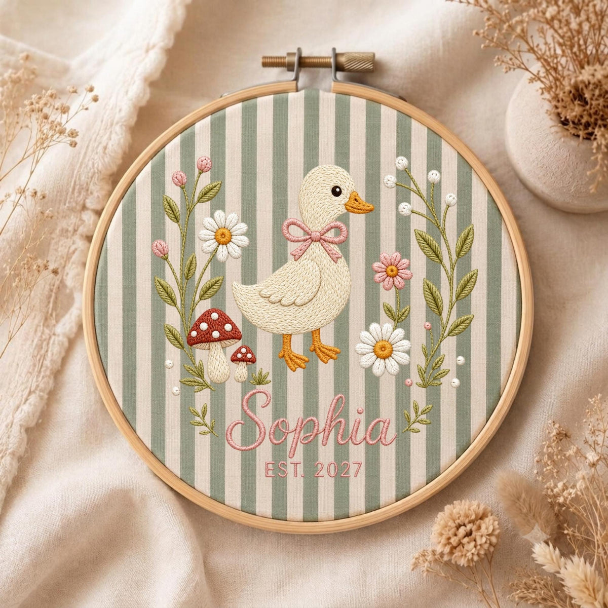 May include: A circular wooden embroidery hoop containing fabric with vertical sage green and white stripes. The design features a central cream-colored duckling with a pink bow, surrounded by embroidered flowers, mushrooms, and the name Sophia Est. 2027.