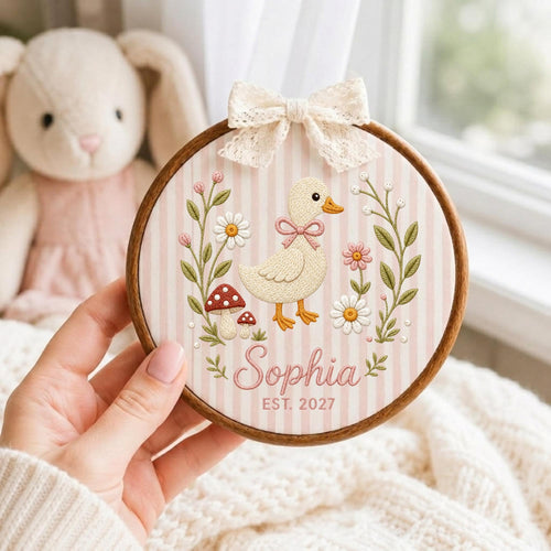 May include: A circular wooden embroidery hoop featuring a light pink and white striped fabric background. The design includes an embroidered cream-colored duck, flowers, and mushrooms, with the name Sophia and EST. 2027, topped with a cream lace bow.