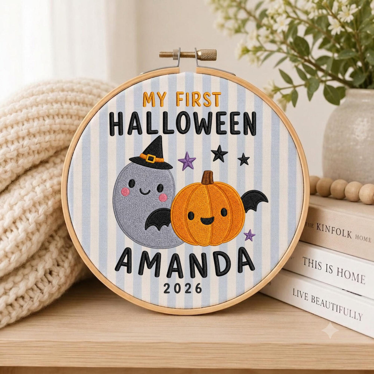 May include: A circular embroidery hoop featuring a light blue and white striped fabric background. The design includes the embroidered text MY FIRST HALLOWEEN AMANDA 2026, a grey ghost wearing a black witch hat, and an orange pumpkin with black wings.