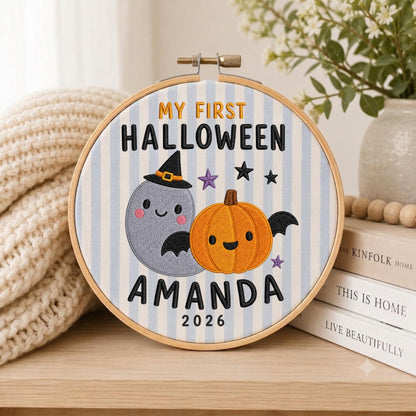 May include: A circular embroidery hoop featuring a light blue and white striped fabric background. The design includes the embroidered text MY FIRST HALLOWEEN AMANDA 2026, a grey ghost wearing a black witch hat, and an orange pumpkin with black wings.