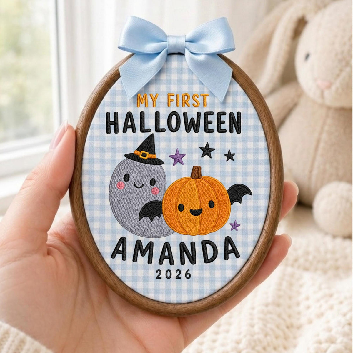 May include: An oval wooden embroidery hoop containing fabric with a light blue gingham pattern. It features embroidered text reading MY FIRST HALLOWEEN AMANDA 2026, a grey ghost wearing a witch hat, and an orange pumpkin with black bat wings.