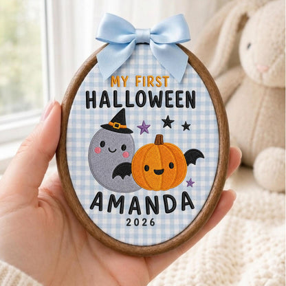 May include: An oval wooden embroidery hoop containing fabric with a light blue gingham pattern. It features embroidered text reading MY FIRST HALLOWEEN AMANDA 2026, a grey ghost wearing a witch hat, and an orange pumpkin with black bat wings.