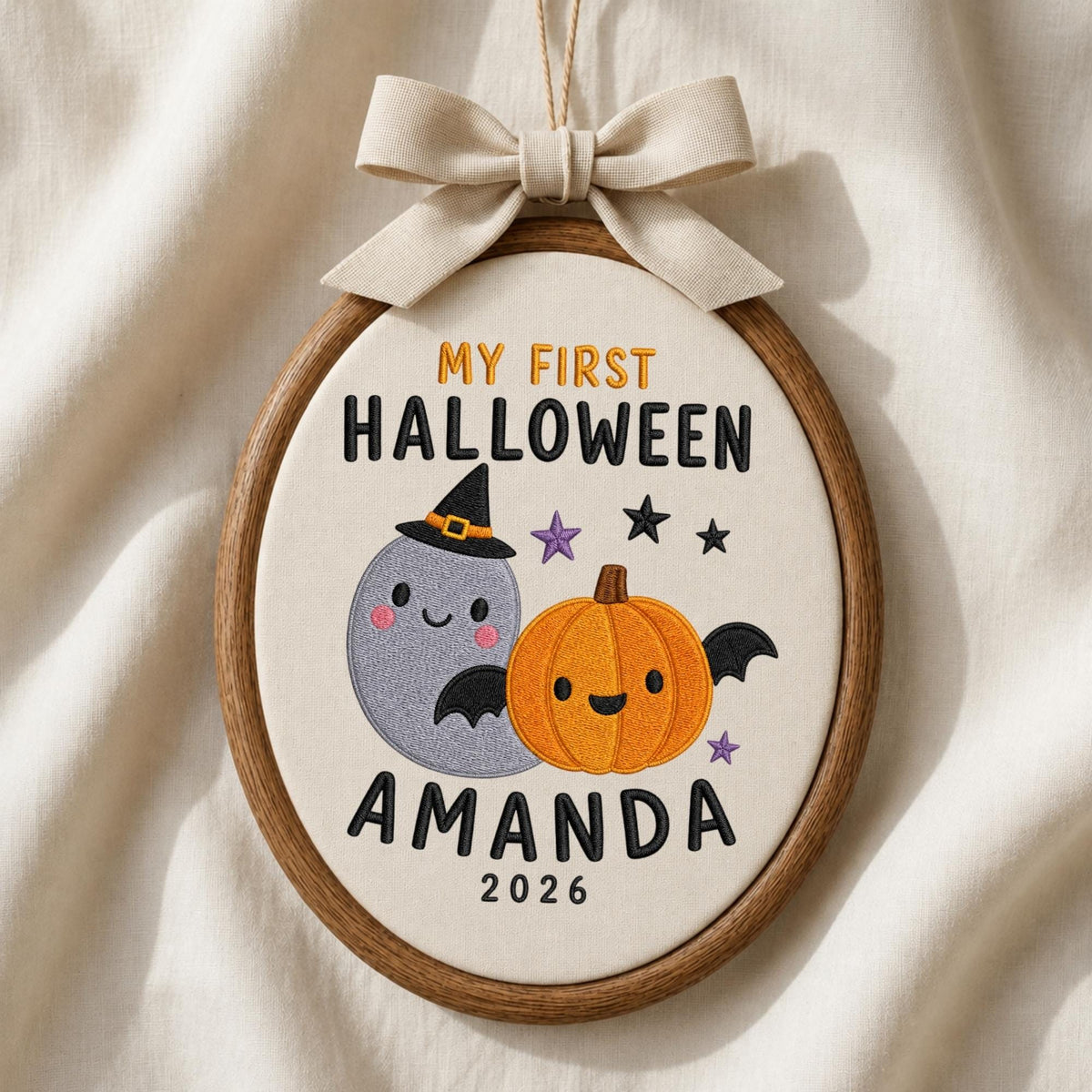 May include: An oval embroidery hoop containing off-white fabric with the embroidered text MY FIRST HALLOWEEN AMANDA 2026. The design features a grey ghost wearing a black witch hat and an orange pumpkin with black bat wings and small stars.