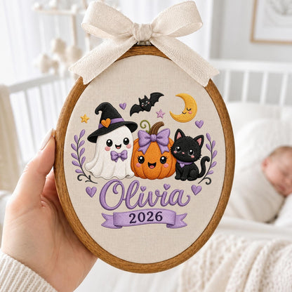 May include: An oval embroidery hoop featuring a cream fabric background with a colorful Halloween-themed design. The illustration includes a white ghost wearing a witch hat, an orange pumpkin with a purple bow, a black cat, a bat, and a yellow crescent moon. The name Olivia and the year 2026 are embroidered in purple. A beige ribbon bow is attached to the top.