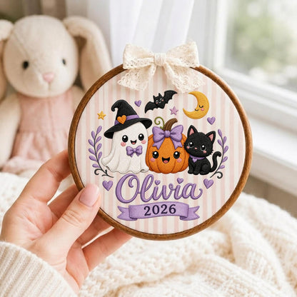 May include: A round wooden embroidery hoop featuring a fabric background with light pink vertical stripes. The design includes an embroidered white ghost wearing a witch hat, an orange pumpkin with a purple bow, and a black cat. The text reads Olivia 2026. A cream lace bow is attached to the top.