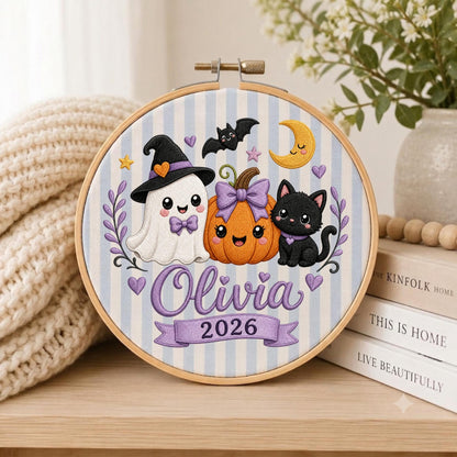 May include: An embroidery hoop featuring a light blue and white striped fabric with a ghost wearing a witch hat, an orange pumpkin with a purple bow, and a black cat. The design includes a yellow crescent moon, stars, hearts, and the text Olivia 2026.