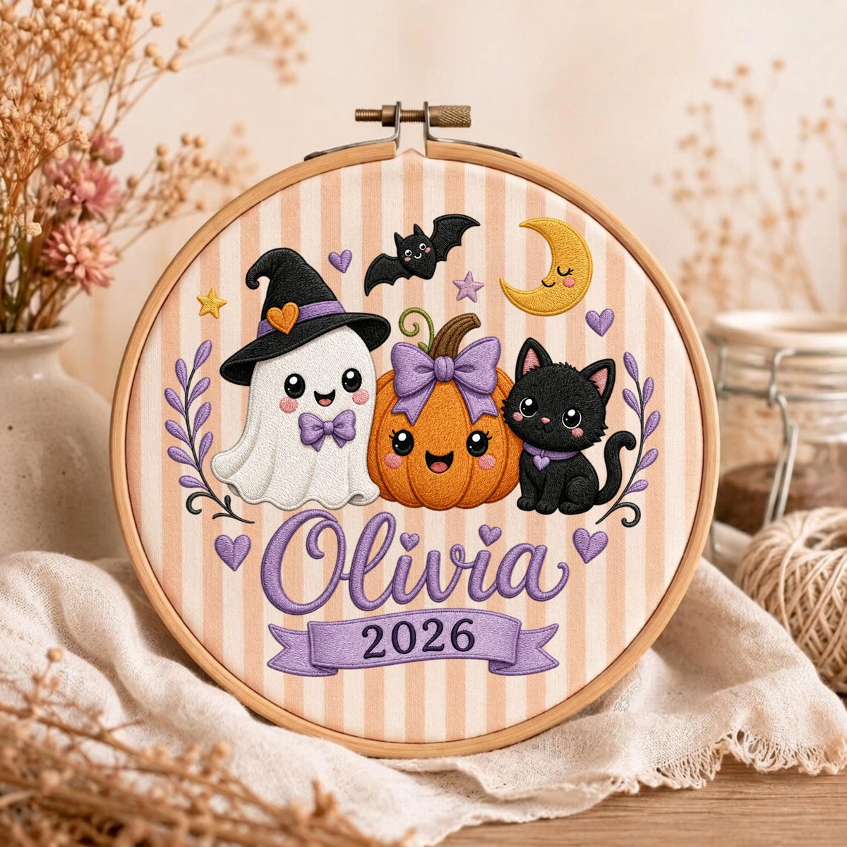 May include: A round wooden embroidery hoop featuring a fabric background with vertical beige stripes. The design includes an embroidered white ghost wearing a witch hat, an orange pumpkin with a purple bow, and a black cat. Text reads Olivia 2026.