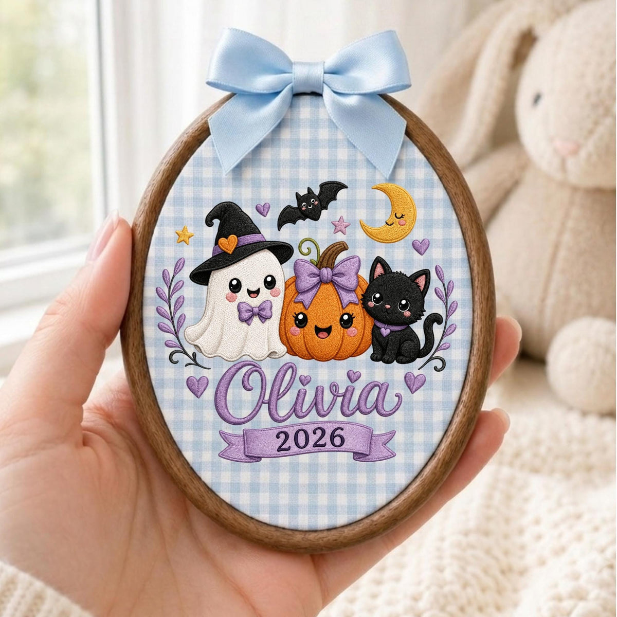 May include: An oval wooden embroidery hoop ornament featuring a light blue gingham fabric background. The design includes a white ghost in a witch hat, an orange pumpkin with a purple bow, and a black cat. Text reads Olivia and 2026.
