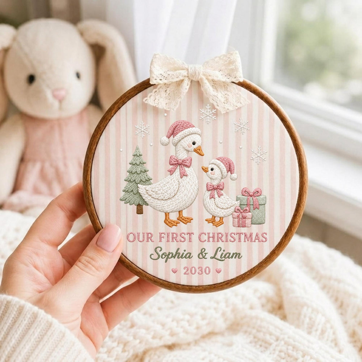May include: A round wooden embroidery hoop featuring a light pink and white striped fabric background. The design shows two white geese wearing pink Santa hats and bows, a green Christmas tree, gifts, and snowflakes. Text reads: OUR FIRST CHRISTMAS Sophia &amp; Liam 2030. A cream lace bow is attached to the top.