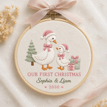 May include: A circular wooden embroidery hoop containing fabric with a stitched design of two white geese wearing pink Santa hats and bows. The scene includes a green pine tree, two gift boxes, and snowflakes. Text reads: OUR FIRST CHRISTMAS Sophia &amp; Liam 2030.