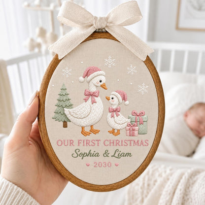 May include: An oval wooden embroidery hoop containing fabric with an embroidered scene of two geese wearing pink hats and bows, a green tree, and gifts. The text reads "OUR FIRST CHRISTMAS", "Sophia &amp; Liam", and "2030". A beige ribbon bow is attached to the top.