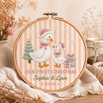 May include: A round wooden embroidery hoop featuring a beige and white striped fabric background. The design shows two white geese wearing pink Santa hats and bows, a green pine tree, snowflakes, and two gift boxes. Text reads: OUR FIRST CHRISTMAS Sophia &amp; Liam 2030.