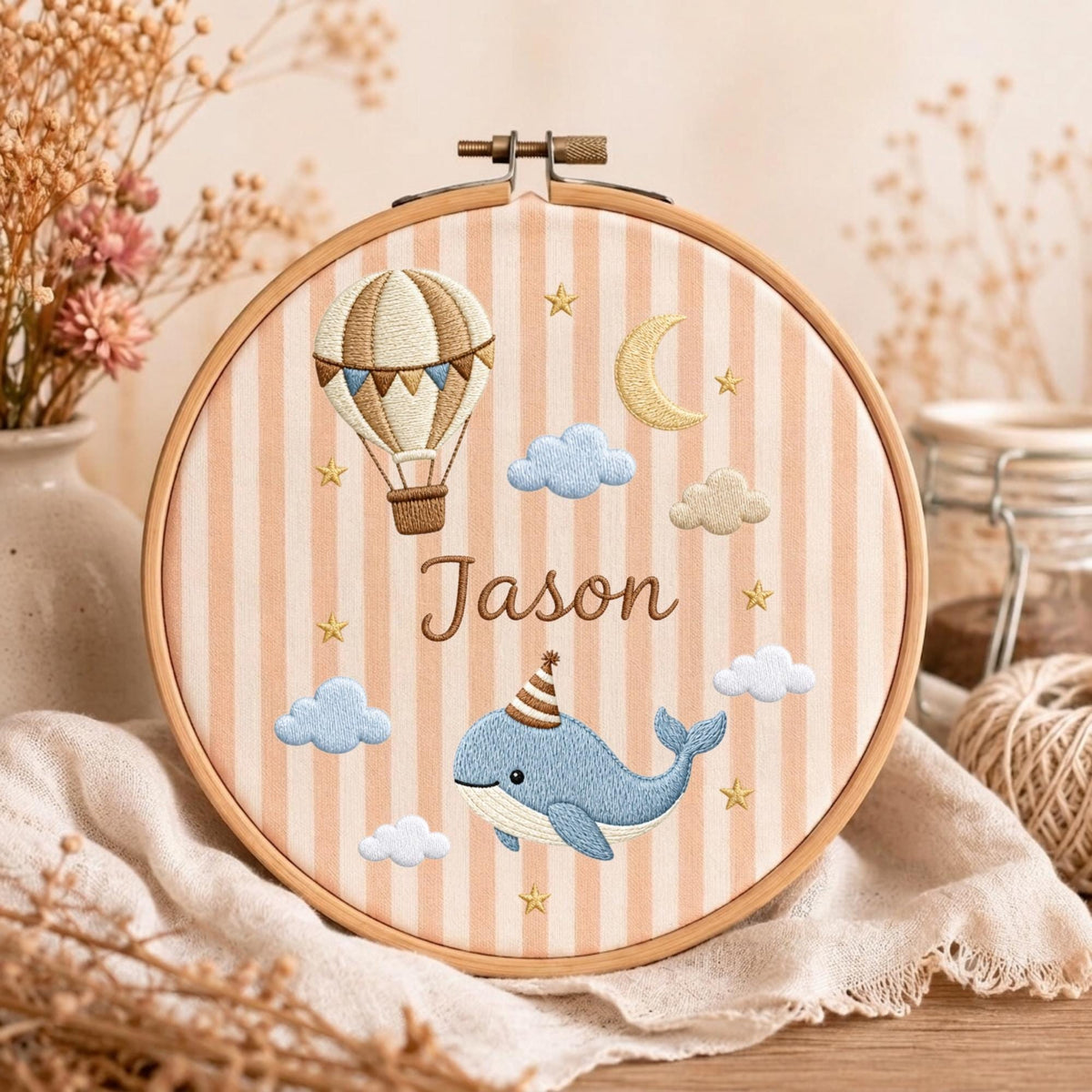 May include: A round wooden embroidery hoop containing fabric with vertical peach and white stripes. The embroidery features a hot air balloon, a yellow crescent moon, blue and white clouds, gold stars, and a blue whale wearing a party hat.