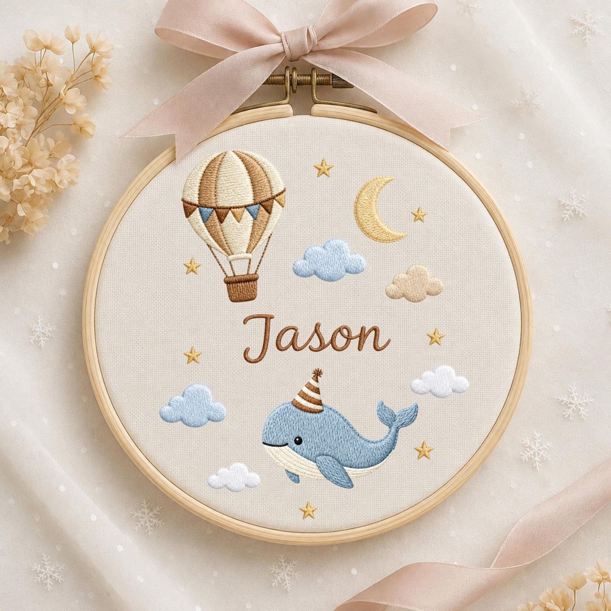 May include: A circular wooden embroidery hoop featuring a beige fabric background with a stitched hot air balloon, a crescent moon, clouds, stars, and a blue whale wearing a party hat. The name Jason is embroidered in brown script in the center.