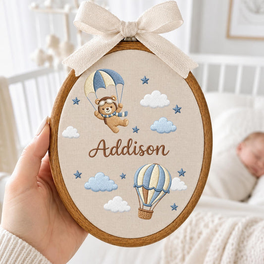 May include: An oval wooden embroidery hoop containing beige fabric with an embroidered brown bear wearing goggles and a scarf, suspended by a blue and cream parachute. Below, a blue and cream hot air balloon, clouds, and stars are stitched, with the name Addison in brown script. A cream ribbon bow is tied at the top.