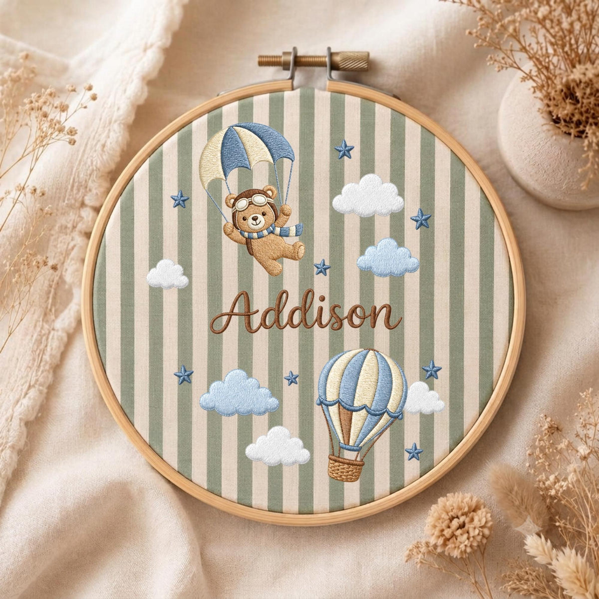 May include: A circular wooden embroidery hoop containing fabric with vertical sage green and cream stripes. The fabric features embroidered designs of a teddy bear with a parachute, a hot air balloon, clouds, blue stars, and the name Addison.