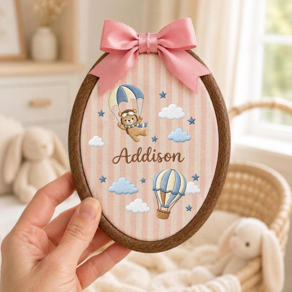 May include: An oval wooden embroidery hoop frame containing fabric with vertical pink stripes. The fabric features embroidered blue and white clouds, blue stars, a hot air balloon, and a bear in a parachute. The name Addison is stitched in brown.