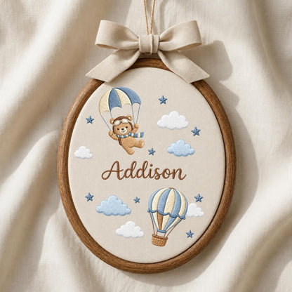 May include: An oval embroidery hoop featuring a beige fabric background with a brown bear wearing aviator goggles and a scarf, suspended by a blue and beige parachute. Below, a blue and beige hot air balloon, clouds, stars, and the name Addison.