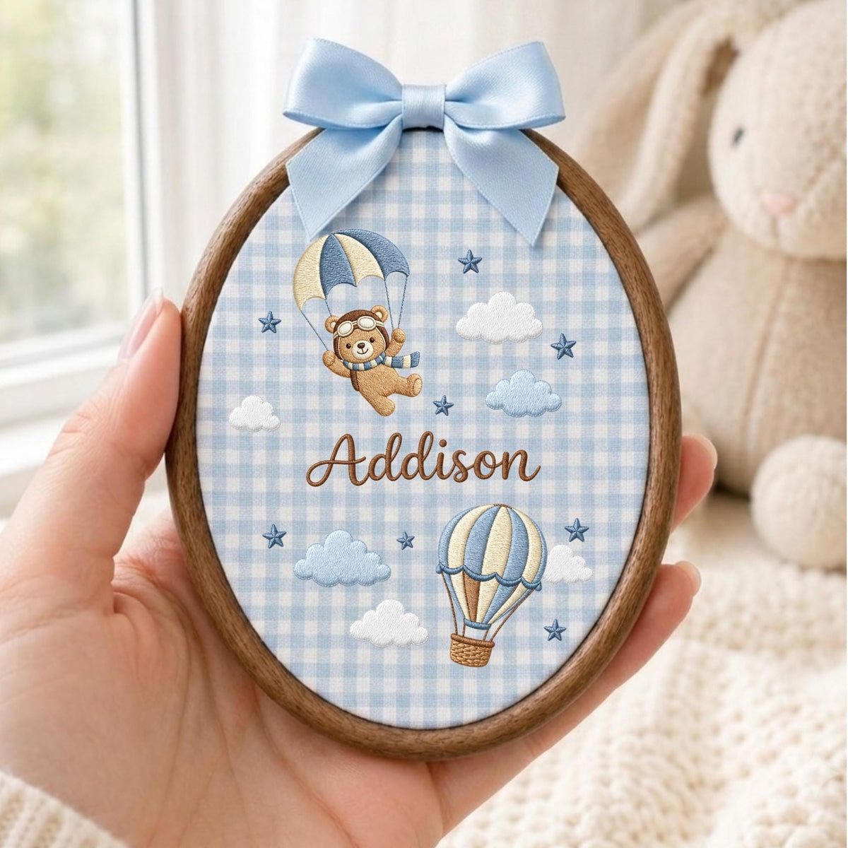May include: An oval wooden embroidery hoop featuring light blue and white gingham fabric. The design includes an embroidered brown bear in a parachute, a hot air balloon, clouds, stars, and the name Addison in brown script with a blue satin bow.