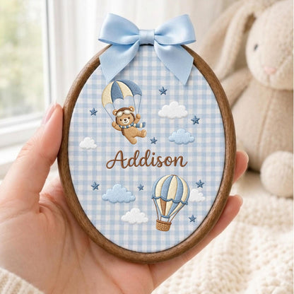 May include: An oval wooden embroidery hoop featuring light blue and white gingham fabric. The design includes an embroidered brown bear in a parachute, a hot air balloon, clouds, stars, and the name Addison in brown script with a blue satin bow.