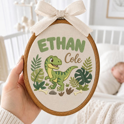 May include: An oval wooden embroidery hoop containing a beige fabric panel with the embroidered green text ETHAN and brown script text Cole. A green dinosaur is centered among embroidered green tropical leaves, bones, and rocks, topped with a bow.