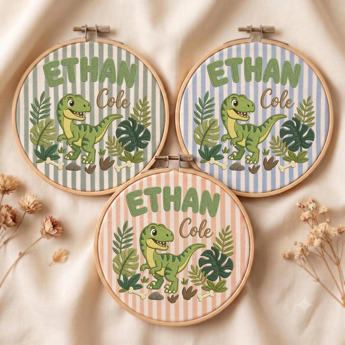 May include: Three circular wooden embroidery hoops featuring fabric with vertical stripes in sage green, light blue, and peach. Each hoop displays an embroidered green dinosaur, tropical leaves, bones, and the name Ethan Cole in green and brown text.