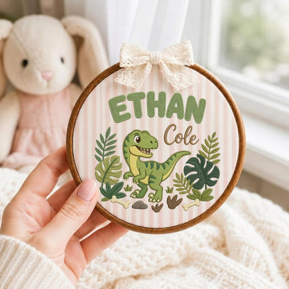 May include: A circular wooden embroidery hoop featuring a light pink and white striped fabric background. The design includes the embroidered green text Ethan Cole, a green dinosaur, various green tropical leaves, and small beige bones.