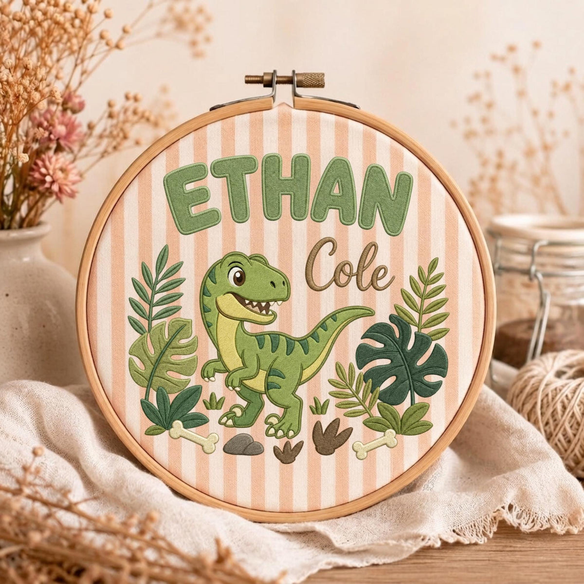 May include: A circular wooden embroidery hoop framing a light pink and white striped fabric. The fabric features an embroidered green dinosaur, tropical leaves, bones, and the text "ETHAN" in block letters and "Cole" in script lettering.